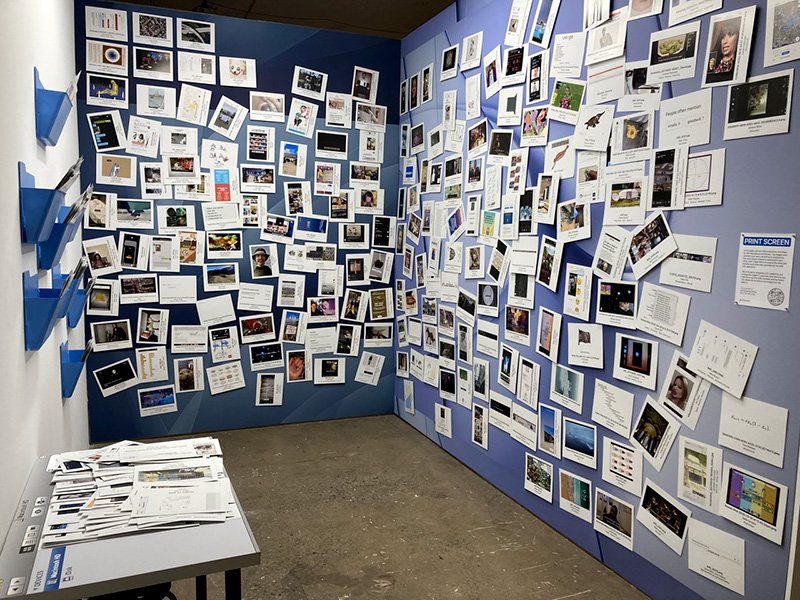 a slim gallery with three visible walls, two are plastered with a giant representation of a standard Mac desktop background which is covered in moveable printed screenshots, the other wall has physical folders that can hold the screenshots and a table that looks like a finder window
